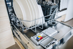 Colorful dishwasher detergent tablet for dishwashing machine. Open dishwasher with clean glasses and dishes close up.